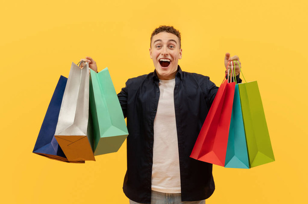 vzrušený mladý muž na sobě ležérní oblečení drží barevné papírové tašky nákupy v obou rukou a křičí, těší nakupování během sezóny prodej, žluté studio pozadí - Fotografie, Obrázek