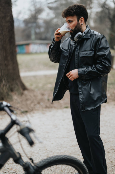 Skupiony biznesmen korzysta z przerwy na kawę na świeżym powietrzu, ubrany w czarną skórzaną kurtkę ze słuchawkami i rower obok niego. - Zdjęcie, obraz