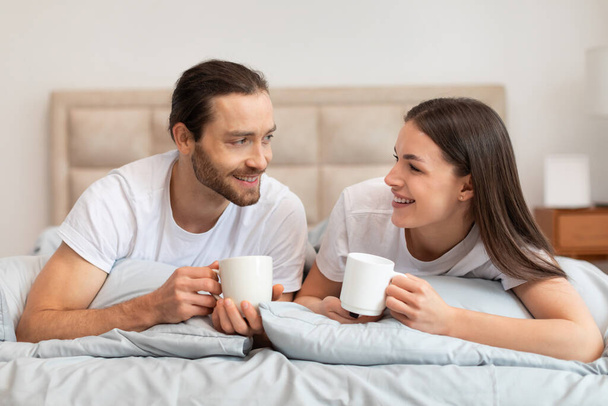 Relaksująca i szczęśliwa para w białych koszulkach, ciesząca się poranną kawą w łóżku, wymieniająca uśmiechy i ciesząca się swoim towarzystwem w spokojnej sypialni - Zdjęcie, obraz