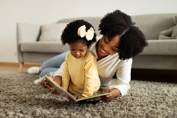 Hora de jugar. Alegre cariñosa madre afroamericana leyendo libro con su hija pequeña, niña señalando imágenes coloridas, pasar tiempo libre juntos en el interior de casa, espacio de copia - Foto, imagen