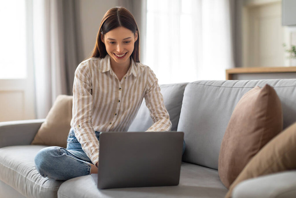 Wesoła młoda kobieta freelancer w luźnej koszuli w paski koncentruje się na laptopie siedząc skrzyżowane nogi na przytulnej kanapie w dobrze oświetlonej przestrzeni życiowej, wolnej przestrzeni - Zdjęcie, obraz
