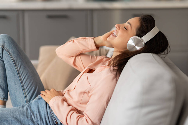 Сяюча жінка лежить на дивані, загублена в музиці з білими навушниками, її обличчя приземляється мирною посмішкою, втілюючи релаксацію та задоволення - Фото, зображення