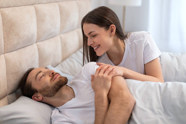 Delikatny moment, gdy uśmiechnięta kobieta z adoracją patrzy na swojego partnera leżącego w łóżku, uchwycając poczucie miłości, opieki i intymności we wnętrzu sypialni - Zdjęcie, obraz