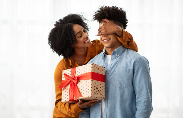 Een aanhankelijke Afro-Amerikaanse duizendjarige vrouw verrast haar partner met een overdekt ooggebaar en een grote geschenkdoos met hartmotief versierd met een rood lint, die liefde en geluk uitstraalt. - Foto, afbeelding