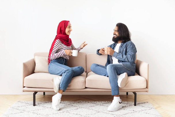 Vrolijk jong moslim paar rust samen thuis, het drinken van koffie en chatten tijdens het zitten op comfortabele bank in de woonkamer, gelukkige islamitische echtgenoten genieten van binnenlandse vrije tijd, vrije ruimte - Foto, afbeelding