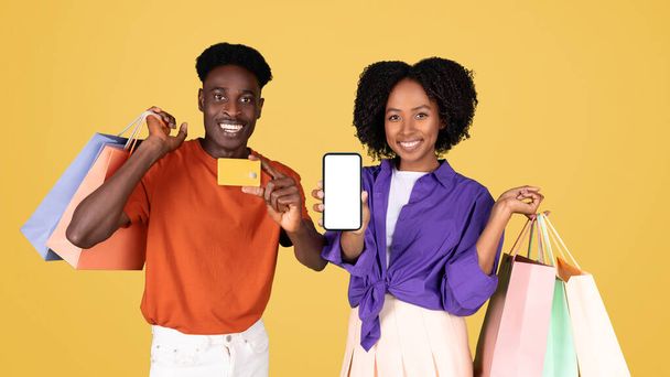 Una coppia vivace gode di una furia di shopping, con l'uomo con una camicia arancione in possesso di una carta di credito e la donna in un top viola sorridente con più borse della spesa su uno sfondo giallo - Foto, immagini
