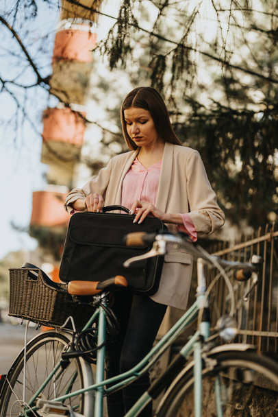 Young businesswoman opening her laptop bag beside a vintage bicycle, urban lifestyle concept - Photo, Image