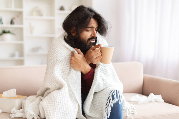 Άρρωστος χιλιετής ανατολικός άντρας έχει κρύωμα ή γρίπη, κάθεται στον καναπέ στο σπίτι, καλυμμένος με κουβέρτα, κρατώντας κούπα, πίνοντας ζεστό τσάι ή φάρμακα, νιώθοντας κρύο - Φωτογραφία, εικόνα