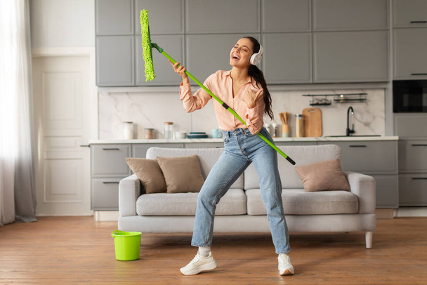 Euphorische Frau mit Kopfhörern benutzt freudig grünen Wischmopp als Mikrofon und singt, während sie ihre helle und stylische Küche putzt und dabei Hausarbeit mit Spaß verbindet - Foto, Bild