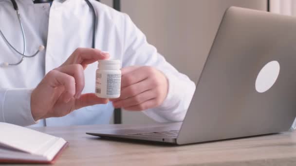 Een arts houdt een fles pillen naast een laptop computer op een tafel, met hun hand duidelijk zichtbaar. - Video