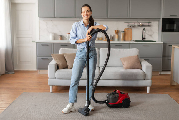Cheerful young woman in casual outfit stands confidently with vacuum cleaner in tidy, well-lit living room, ready for household cleaning, full length - Zdjęcie, obraz