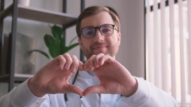 De dokter glimlacht en maakt een hartvorm met zijn handen, toont zijn zorgzame aard in de gezichtsverzorging. - Video