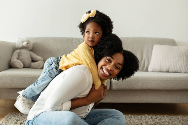Χαρούμενη έννοια της μητρότητας. Αξιολάτρευτη Αφρο-Αμερικάνα με μαλλιαρό φιόγκο να καβαλάει τη γελαστή μαμά της. Χαρούμενη μαύρη μητέρα και κόρη νήπιο να διασκεδάσουν στο σπίτι, πλαϊνή άποψη, αντίγραφο χώρου - Φωτογραφία, εικόνα