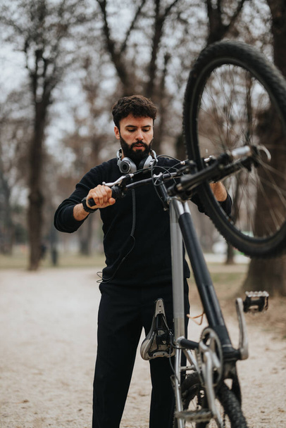 Ένας αφοσιωμένος επιχειρηματίας με casual ενδυμασία προσαρμόζει τα γρανάζια του ποδηλάτου του σε ένα υπαίθριο περιβάλλον, επιδεικνύοντας multitasking και έναν υγιεινό τρόπο ζωής. - Φωτογραφία, εικόνα