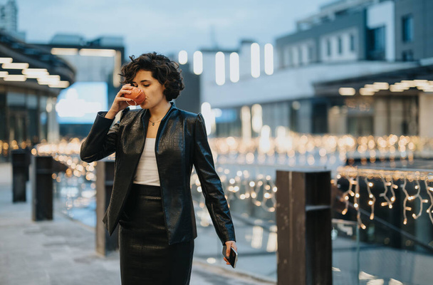 Elegante joven saboreando una bebida caliente mientras está de pie en un paisaje urbano adornado con brillantes luces festivas. - Foto, imagen