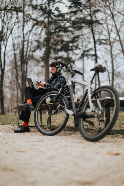 Ein modischer männlicher Unternehmer erledigt geschäftliche Aufgaben auf seinem Laptop in einem ruhigen Park, das Fahrrad an seiner Seite. - Foto, Bild