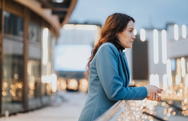 Uma mulher pensativa em um blazer azul se destaca ao ar livre à noite, cercada por luzes quentes da cidade, incorporando contemplação urbana e serenidade. - Foto, Imagem