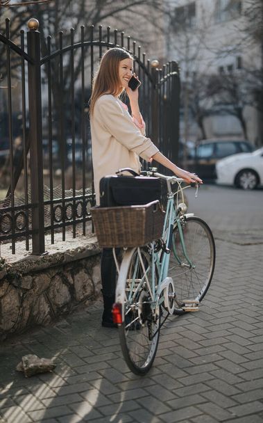 Mutlu genç bir kadın, eski bisikletine yaslanmış, güneşli bir caddede bir sepetle telefon görüşmesi yapıyor.. - Fotoğraf, Görsel