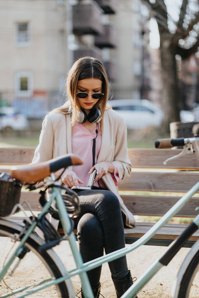Модная молодая женщина проверяет свой смартфон, сидя на скамейке в солнечном парке, с велосипедом рядом. - Фото, изображение