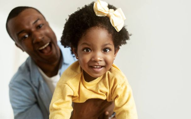 Крупный план портрет милой африканской маленькой девочки с луком в волосах улыбается в камеру, малышка дочь и отец весело провести время вместе дома, белый фон, скопировать пространство - Фото, изображение