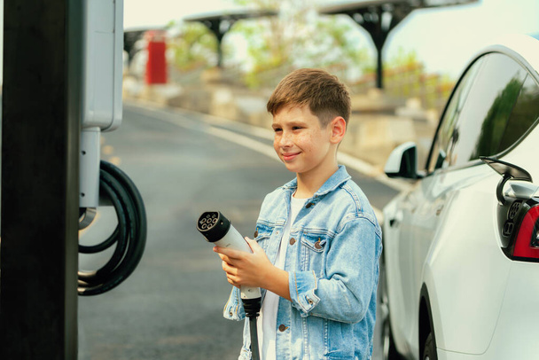 Mały chłopiec ładujący ekologiczny elektryczny samochód ze stacji ładowania EV. Koncepcja podróży samochodem EV dla alternatywnego transportu napędzanego czystą energią odnawialną i zrównoważoną. Wieczne - Zdjęcie, obraz