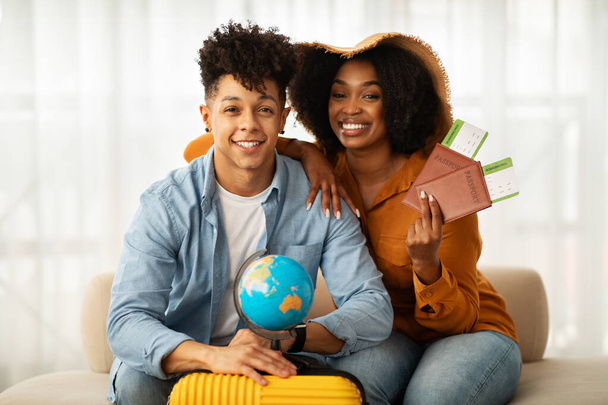 Feliz pareja afroamericana milenaria con pasaportes y tarjetas de embarque listas para viajar, sentadas al lado de un globo y una maleta de color amarillo brillante, irradiando emoción por una nueva aventura - Foto, imagen
