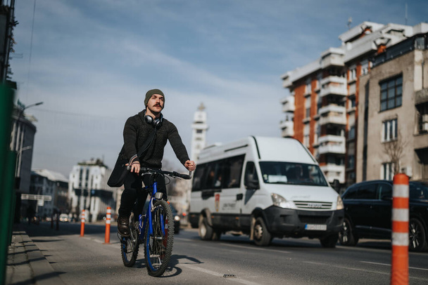 Skupiony mężczyzna dojeżdżający do pracy na rowerze po drodze miejskiej z pojazdami w tle w ciągu dnia. - Zdjęcie, obraz