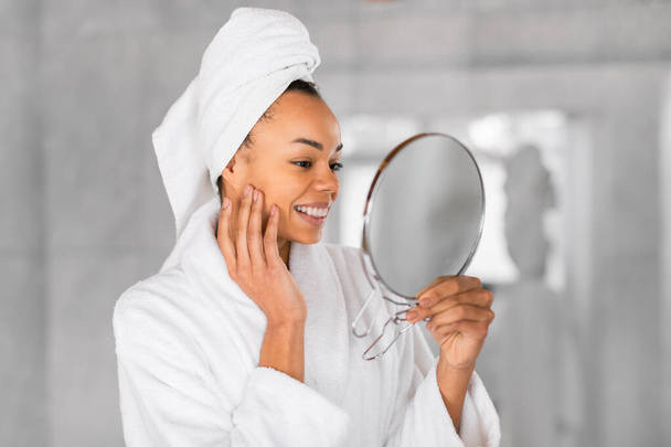 Soins du visage. Jolie femme noire avec une serviette enveloppée sur la tête tient miroir rond, toucher doucement le visage, aspect admiratif avec sourire dans la salle de bain à la maison, portrait shot - Photo, image