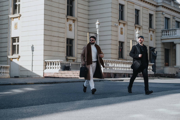 Captura de dois indivíduos masculinos na moda cruzando a estrada com confiança em um dia ensolarado com arquitetura urbana no pano de fundo. - Foto, Imagem