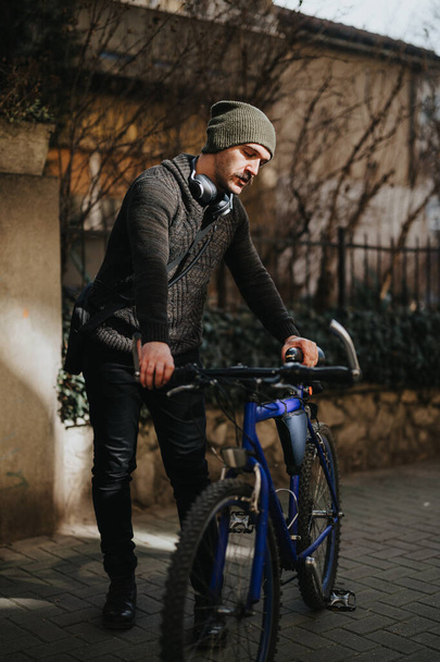 Un uomo elegante in abbigliamento invernale casual si ferma in una strada soleggiata con la sua bicicletta, indossando cuffie intorno al collo. - Foto, immagini