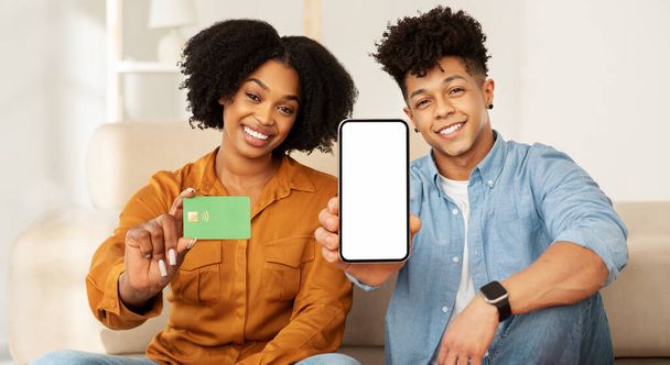 Cheerful african american couple showing a credit card and a smartphone with a blank screen, suggesting online payment or shopping, while comfortably sitting in a well-lit, modern living room - Photo, Image