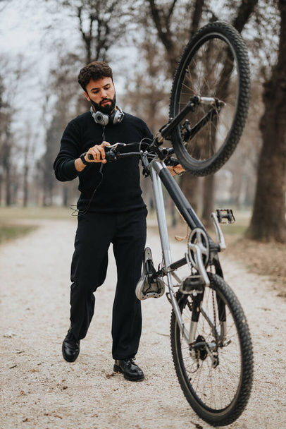 Skupiony biznesmen w formalnym stroju dopracowuje swój rower w spokojnym parku, ze słuchawkami na szyi.. - Zdjęcie, obraz