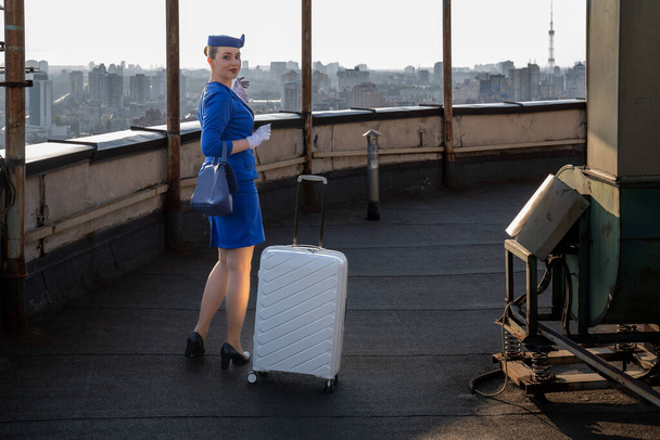 αεροσυνοδός με μπλε vintage στολή με καπέλο, γάντια, βαλίτσα, πόζες τσάντας. Αεροσυνοδός στη χρυσή εποχή των αεροπορικών ταξιδιών. Εργασία στην αεροπορία. Βιομηχανία εξυπηρέτησης πελατών, ταξίδια με αεροσκάφη σε όλο τον κόσμο - Φωτογραφία, εικόνα