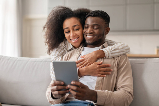 Digitale binding. Liefdevolle duizendjarige Afro-Amerikaanse echtpaar geniet van websurfen op tablet computer, vrouw knuffelen echtgenoot terwijl hij met behulp van gadget, verbetering van hun moderne levensstijl binnen thuis - Foto, afbeelding