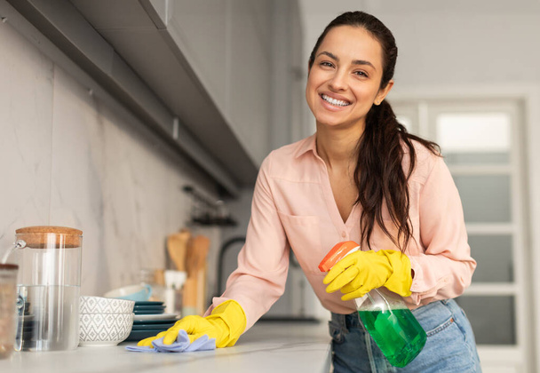 Barátságos nő sugárzó mosollyal, sárga kesztyűt visel, kék ruhával és zöld spray-vel takarítja a konyhaasztalát, élvezi a háztartási rutint - Fotó, kép