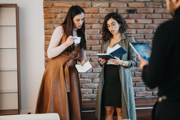 Two professional women discussing work with notepad and coffee in a brick office setting, exuding focused teamwork. - Photo, Image