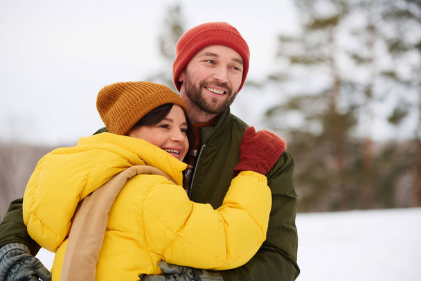Μέτριο πλάνο του χαρούμενου Καυκάσιου ζευγαριού που περνά τη χειμερινή μέρα κάνοντας πεζοπορία στα βουνά, αγκαλιάζοντας και κοιτάζοντας μακριά - Φωτογραφία, εικόνα