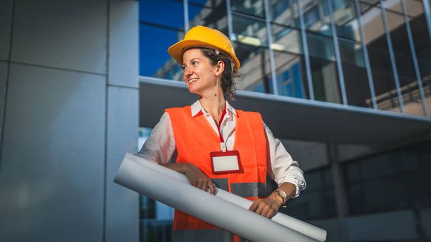 architekt kobieta inżynier budownictwa stoją na zewnątrz nosić kask ochronny i zachód przed nowoczesnym budynku ściany trzymać planów budowlanych - Zdjęcie, obraz