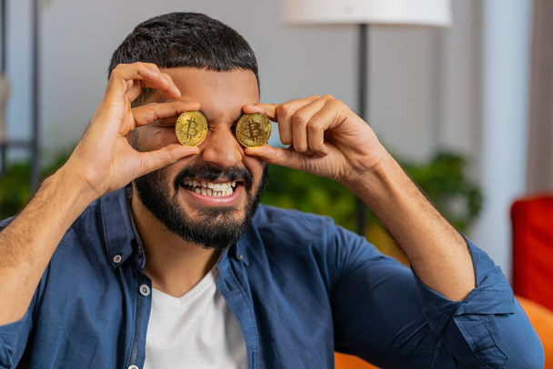 Hymyilevä intialainen freelancer mies varastossa kauppias tilalla kaksi kultaista bitcoineja BTC kattaa silmät. Happy Hispanic mies kaveri onnistunut kehittäjä ohjelmoija istuu sohvalla kotona olohuoneessa. Vaurauden lisääminen. - Valokuva, kuva