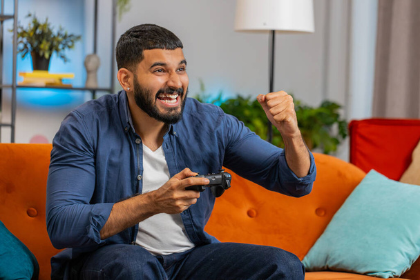 Χαρούμενος νεαρός Ινδός που χρησιμοποιεί χειριστήριο χειριστηρίου που παίζει βιντεοπαιχνίδι και κάθεται στον καναπέ στο σαλόνι. Ισπανόφωνος τύπος σφίγγοντας γροθιές απολαμβάνοντας την επιτυχία κερδίζοντας online παιχνίδι κατά τη διάρκεια του Σαββατοκύριακου στο σπίτι. - Φωτογραφία, εικόνα