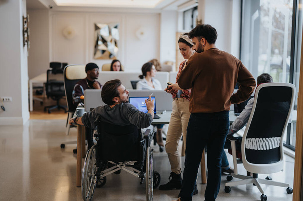 Uma cena de escritório moderna captura uma equipe dinâmica, incluindo um profissional em uma cadeira de rodas discutindo ideias e colaborando em um espaço de trabalho brilhante. - Foto, Imagem