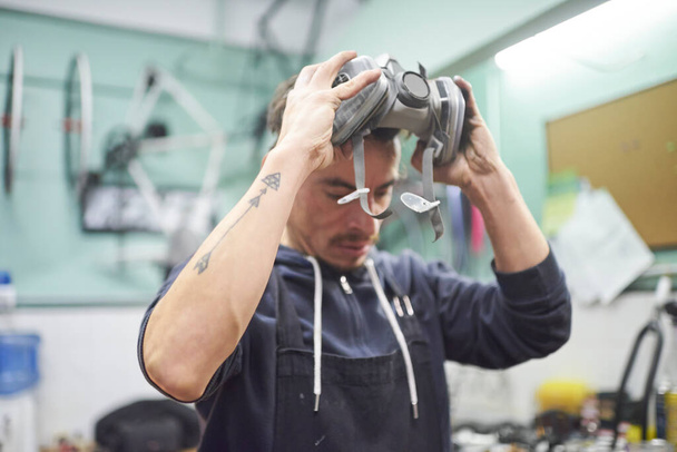 Πορτραίτο ενός ισπανόφωνου νεαρού εργάτη που αφαιρεί μάσκα αναπνευστικής προστασίας, καθώς ολοκληρώνει μια εργασία ζωγραφικής με σπρέι στο εργαστήριό του. Προστασία της υγείας στη βιομηχανική εργασία. Πραγματικοί άνθρωποι εργάζονται. - Φωτογραφία, εικόνα