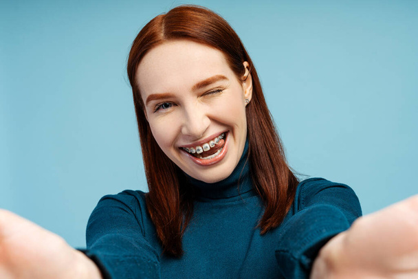Closeup selfie of a joyful redhead woman with braces, wearing a polo neck sweater, capturing herself and looking at the camera, isolated on a blue background - Foto, afbeelding