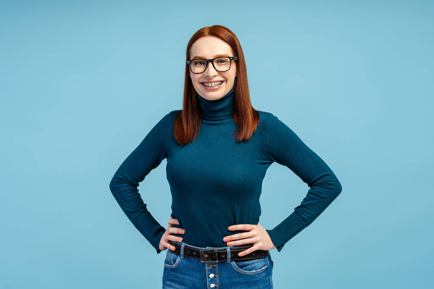 Рыжеволосая женщина в очках и брекетах, в свитере для шеи в поло, кладет руки на бедра, предлагая тёплую улыбку в камеру, изолированную на синем фоне. Концепция умной женщины - Фото, изображение