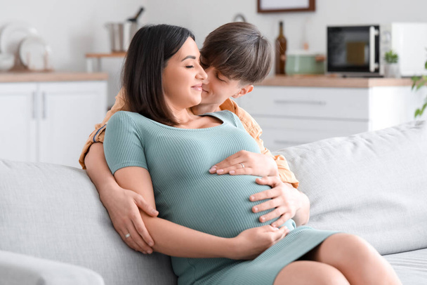 Genç hamile çift evdeki kanepede sarılıyor. - Fotoğraf, Görsel