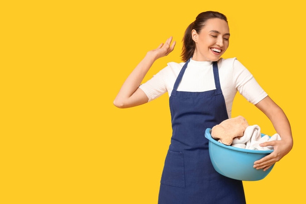Portrait of happy young housewife in apron with dirty laundry on yellow background - Photo, Image