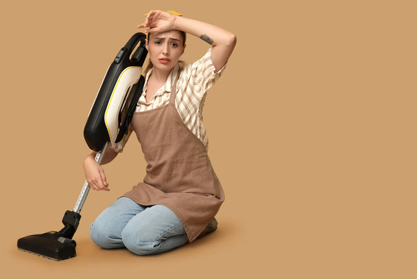 Portrait of tired young housewife in apron with vacuum cleaner on brown background - Photo, Image
