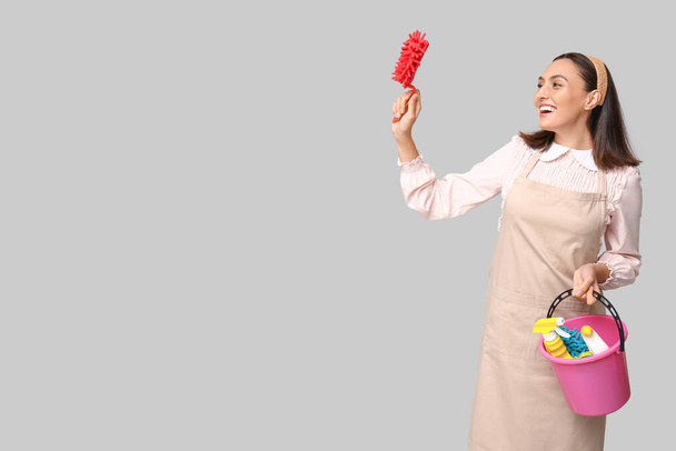Portrait of happy young housewife in apron with cleaning supplies on grey background - Photo, Image