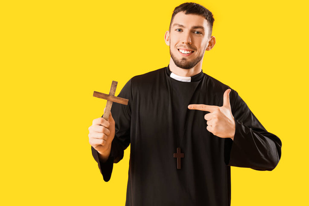 Junger Priester zeigt auf Kreuz auf gelbem Hintergrund - Foto, Bild