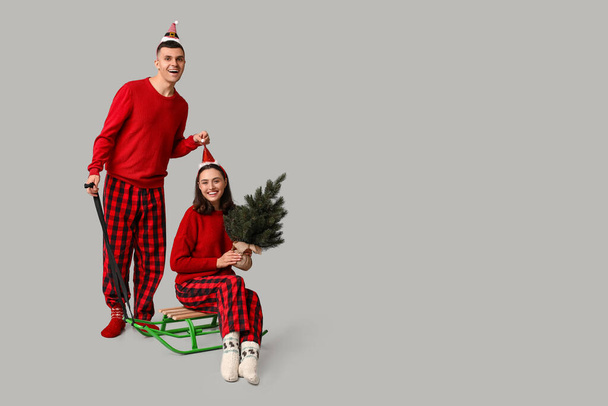 Happy young couple in Christmas pajamas, with sledge and coniferous tree on grey background - Zdjęcie, obraz